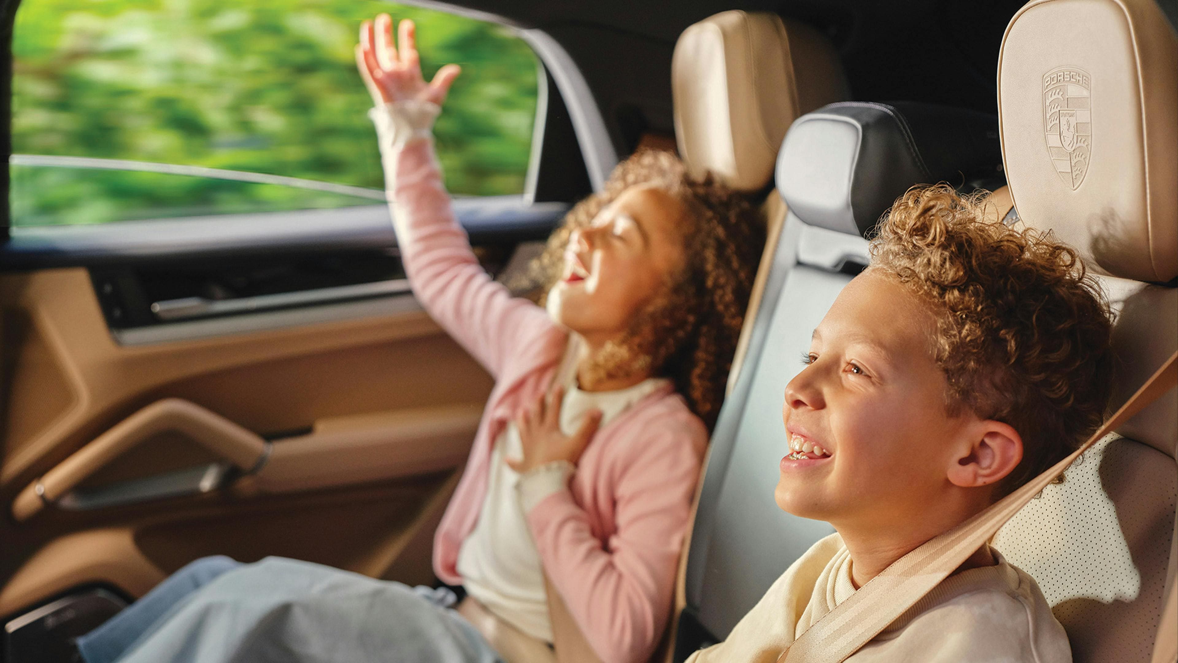 Two children smiling and enjoying a car ride in the backseat of a Porsche.